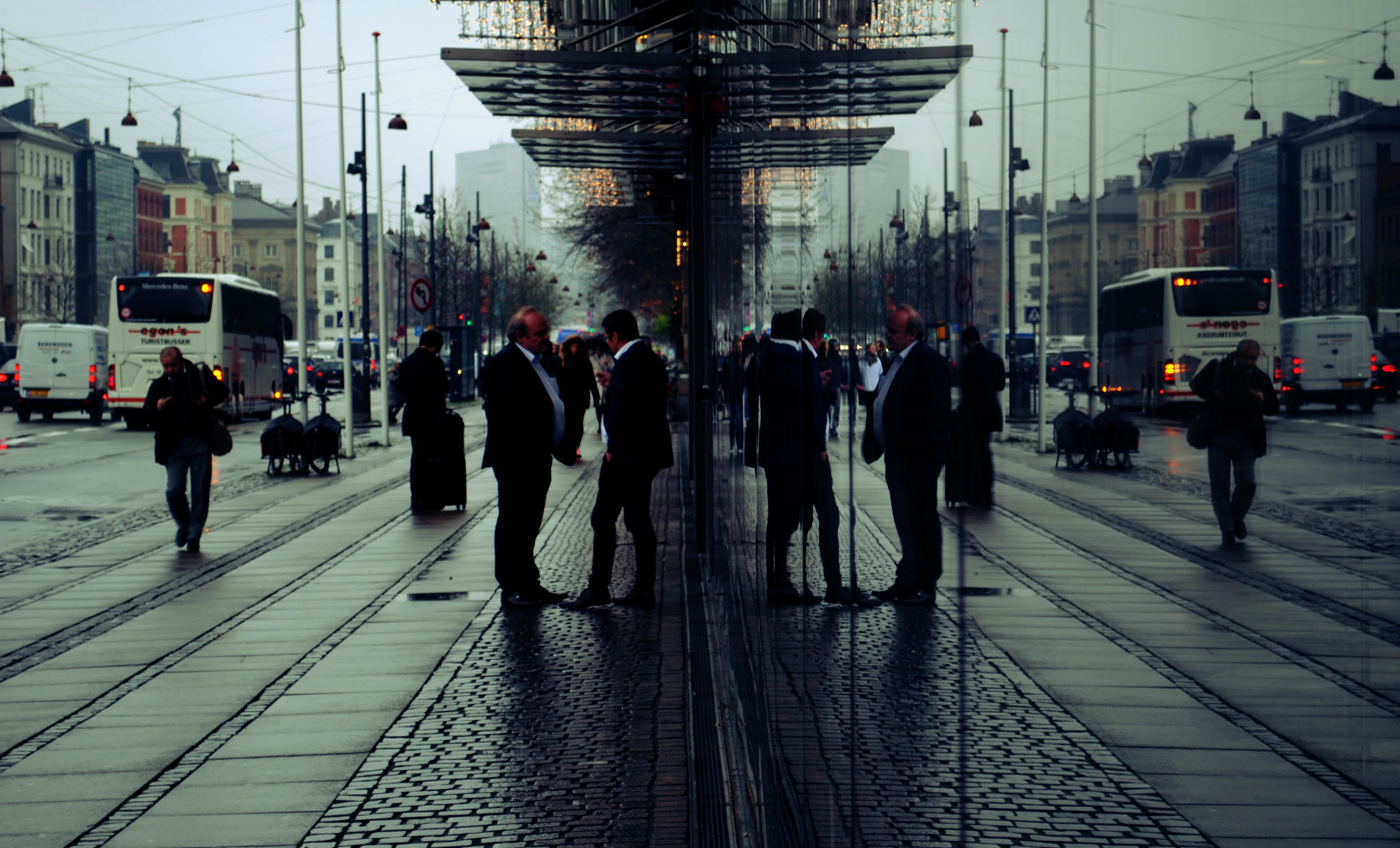 Two men in suits talking at a tram stop, reflections in a mirrored wall