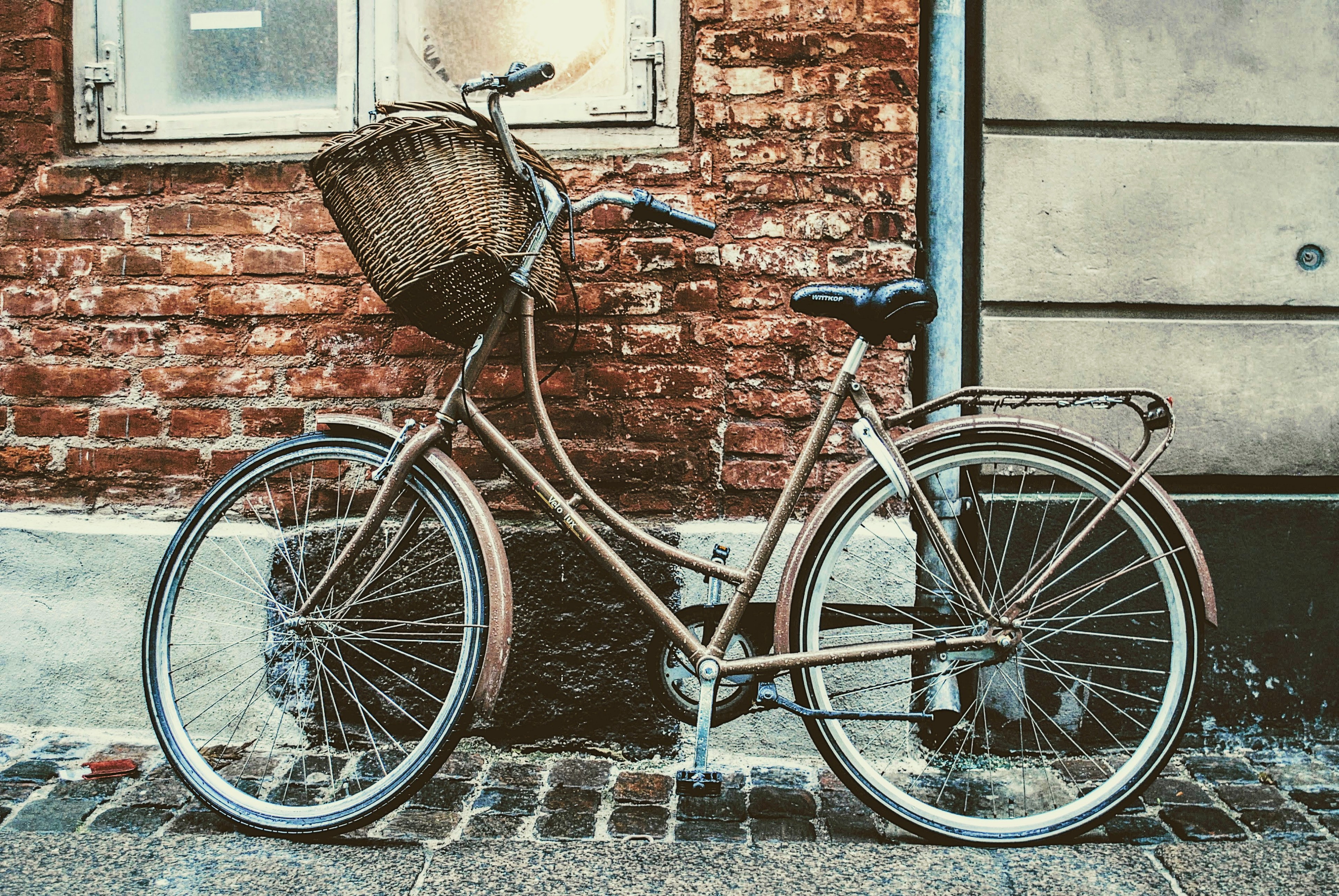 A rust-brown city bike with a wicker basket against a red brick wall