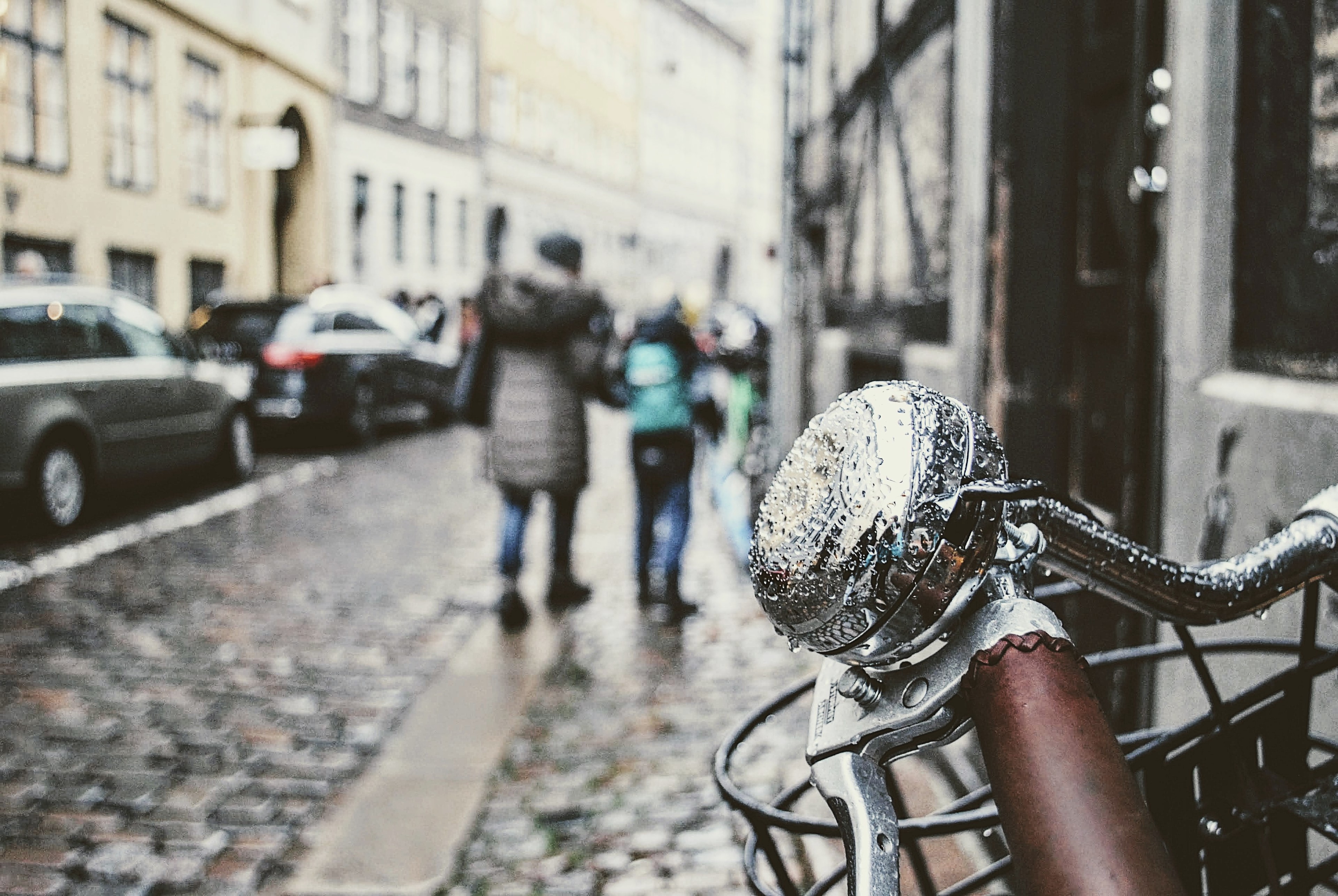 A bicycle headlight beaded with rain, figures walking a cobblestone street